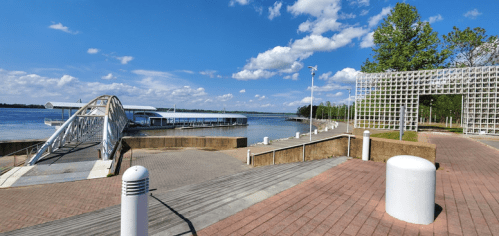 A scenic riverside view featuring a dock, pathway, and greenery under a blue sky with scattered clouds.