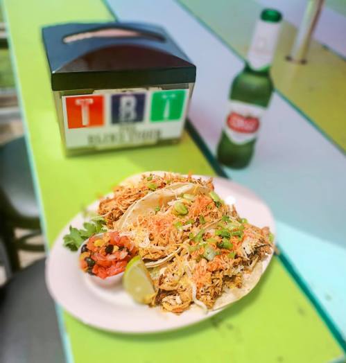A plate of three tacos topped with shredded meat, lettuce, and salsa, alongside a lime wedge and a beer bottle.