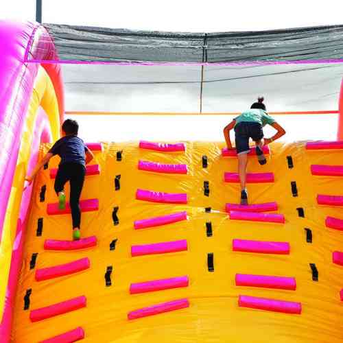Two children climbing a bright yellow inflatable wall with pink grips under a sunny sky.