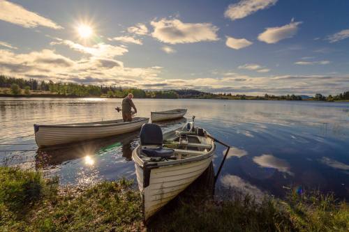 A person stands by a calm lake with two boats, reflecting clouds and sunlight in the water.