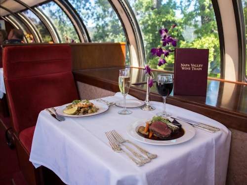 A dining table set with two gourmet dishes, wine glasses, and a menu in a scenic train car with large windows.