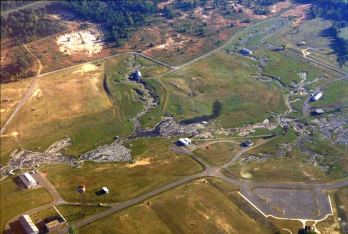 Aerial view of a landscape with scattered buildings, winding paths, and patches of grass, showing signs of land alteration.