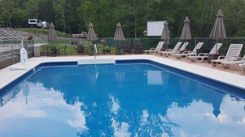 A clear blue swimming pool surrounded by lounge chairs and umbrellas, set in a green, wooded area.