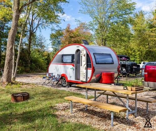 A small, modern camper trailer parked in a wooded area, with picnic tables and chairs nearby.