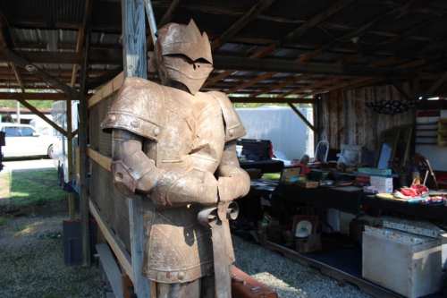 A rusted metal knight statue stands in a rustic shed filled with various items and tools.