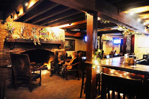 Cozy interior of a bar with a fireplace, wooden beams, and patrons seated near the hearth, decorated for a festive atmosphere.
