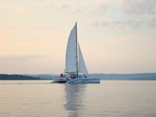 A sailboat with white sails glides across calm waters during sunset, surrounded by distant hills.
