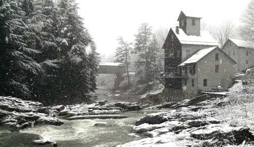 A snowy landscape featuring a river, rocky shore, and a historic building surrounded by trees.