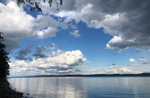 A serene lake scene with calm waters reflecting a blue sky filled with fluffy clouds and distant hills.