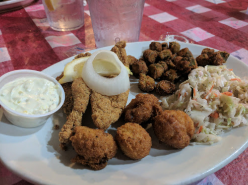A plate of fried fish, onion rings, hushpuppies, coleslaw, and fried okra, served with a side of dipping sauce.