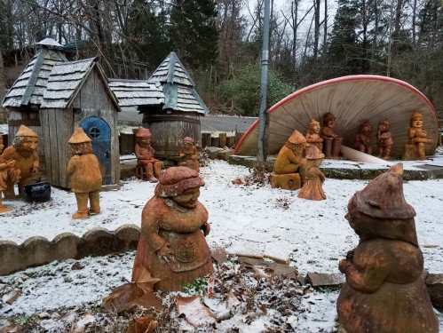 A whimsical garden scene featuring clay gnomes, a small wooden house, and a large shell sculpture, all in a snowy setting.