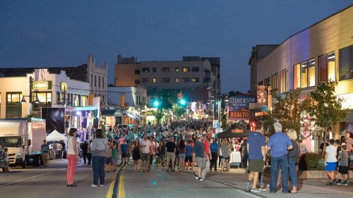 A bustling street scene at night, filled with people walking among shops and food stalls under bright lights.