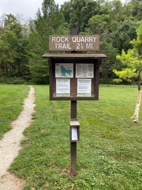 Sign for Rock Quarry Trail, 2.25 miles, with trail map and information, surrounded by greenery and a gravel path.