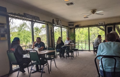 A cozy restaurant interior with diners at tables, large windows showing greenery outside, and a welcoming atmosphere.