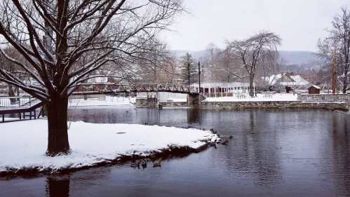 A snowy landscape featuring a tree, a bridge, and a calm river, with a quaint building in the background.