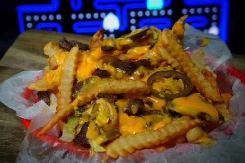 A close-up of loaded fries topped with cheese, jalapeños, and beef, served in a red basket against a retro gaming backdrop.