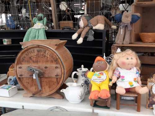 A vintage display featuring a wooden churn, dolls, and a teapot on a table at an antique market.