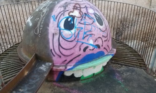 A colorful, graffiti-covered dome with a slide, resembling a cartoonish face, set in a grassy area.