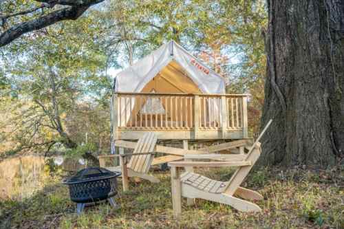 A cozy glamping tent on a wooden deck by a pond, with two Adirondack chairs and a fire pit nearby.