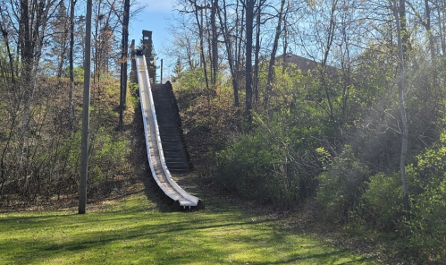 A tall, empty waterslide descends from a wooded hill, surrounded by greenery and sunlight.