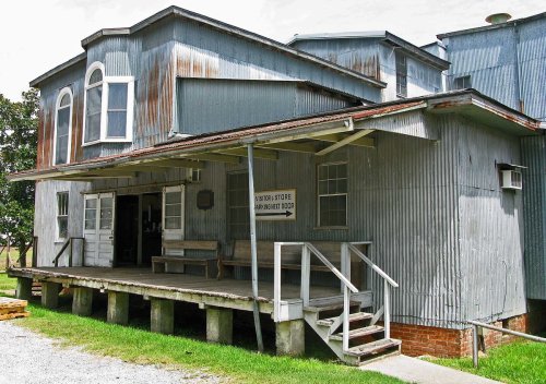 A weathered, gray metal building with a porch and steps, labeled as a "General Store," surrounded by grass.