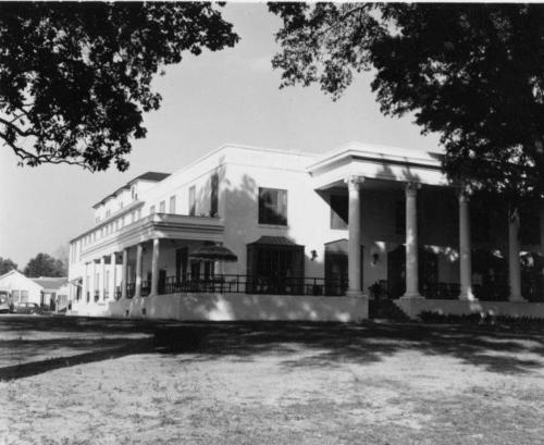 A large, white, two-story building with columns and a patio, surrounded by trees and a grassy area. Black and white photo.