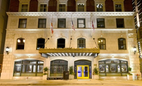 Historic building facade illuminated at night, featuring large windows, flags, and a bright yellow entrance.