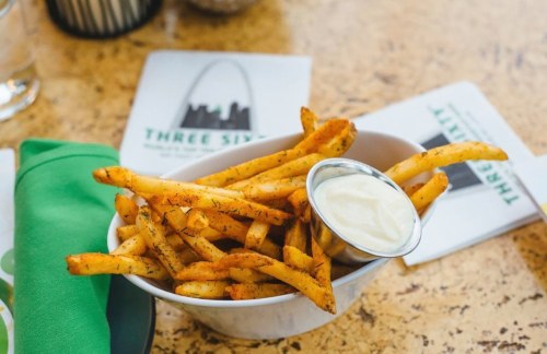 A bowl of seasoned French fries with a small cup of dipping sauce, placed on a textured table with menus nearby.