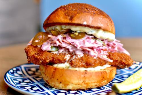 A crispy fried chicken sandwich with coleslaw and jalapeños on a toasted bun, served on a decorative plate.