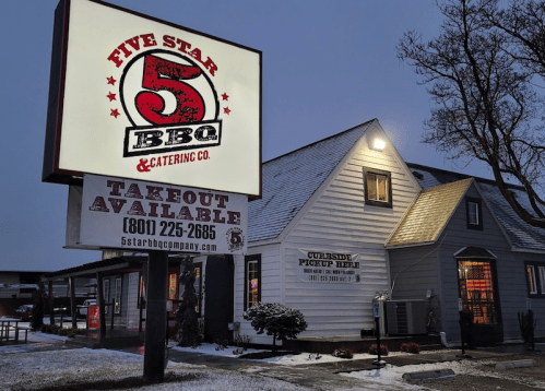 A BBQ restaurant with a lit sign, featuring a takeout option, set in a snowy evening scene.