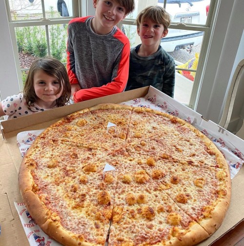 Three children smile beside a large cheese pizza in a box, with a sunny window in the background.
