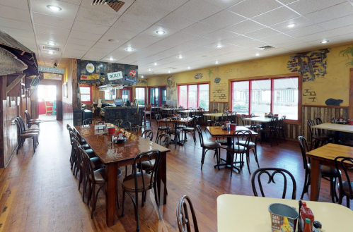 A spacious restaurant interior with wooden tables, chairs, and colorful decor, featuring large windows and a welcoming atmosphere.