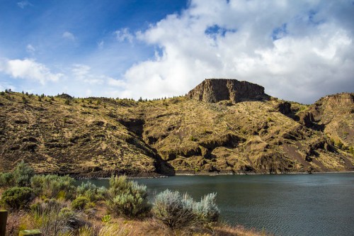 A serene landscape featuring a calm lake, rocky hills, and a cloudy blue sky in the background.