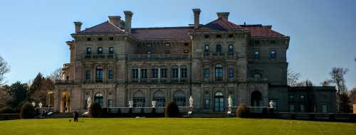 A grand mansion with ornate architecture, surrounded by manicured lawns and trees under a clear blue sky.