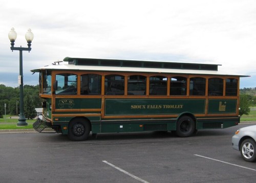 A green and wooden trolley bus parked on a street, with a scenic view in the background.