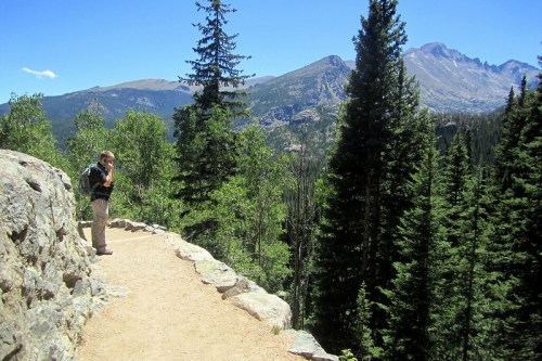 A person stands on a rocky path surrounded by trees, with mountains in the background under a clear blue sky.