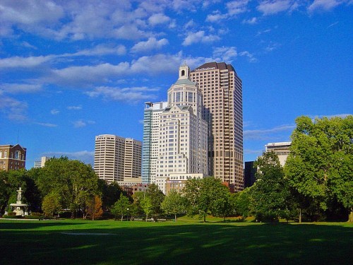 City skyline featuring modern skyscrapers against a blue sky, with green trees and a park in the foreground.