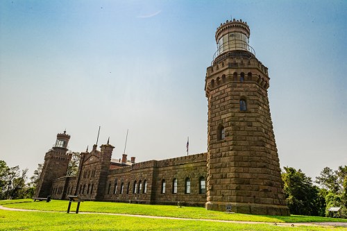 Historic stone lighthouse and building under a clear blue sky, surrounded by green grass and trees.
