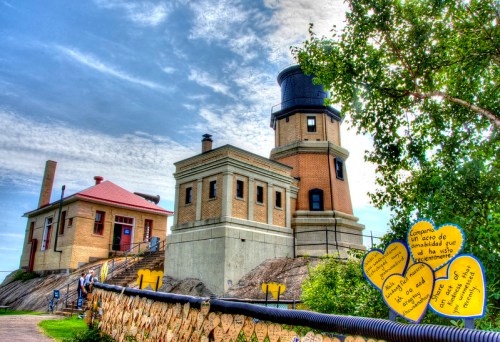 Historic lighthouse with a blue dome, surrounded by greenery and colorful signs, under a partly cloudy sky.