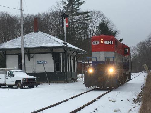 A red and blue train approaches a snowy station with a signal post, surrounded by trees and a parked truck.