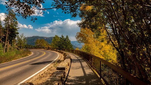 A winding road alongside a wooden walkway, surrounded by trees and vibrant autumn foliage under a blue sky.
