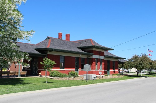 Historic red brick building with a sloped roof, surrounded by greenery and a clear blue sky.