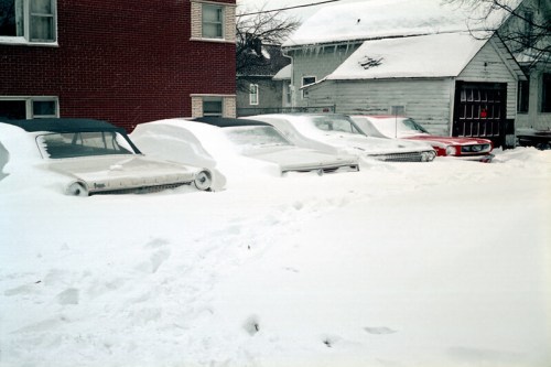 A row of parked cars partially buried in deep snow, with a brick building and a house in the background.