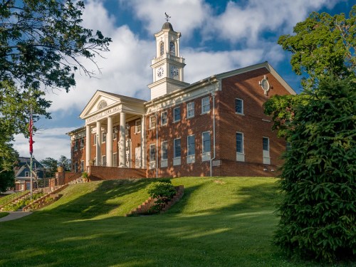 Historic brick building with a clock tower, surrounded by green grass and trees under a partly cloudy sky.