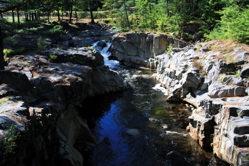 A serene river flows through rocky terrain, surrounded by lush green trees and sunlight filtering through the foliage.