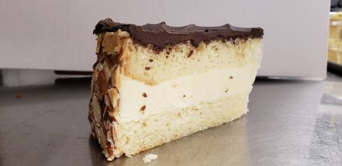 A slice of layered cake with chocolate frosting, creamy filling, and almond topping on a silver surface.