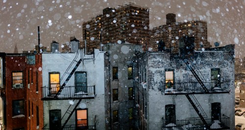 Snow falls over urban buildings at night, with illuminated windows and a soft glow from streetlights.