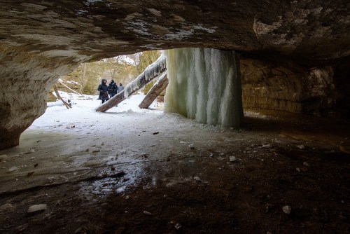 Two people stand near an icy cave entrance, with frozen water formations and a snowy landscape visible outside.