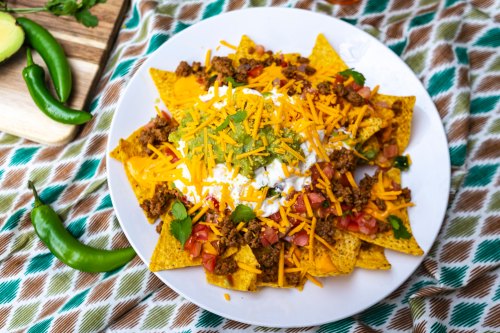 A plate of nachos topped with ground beef, cheese, tomatoes, sour cream, and green sauce, surrounded by green chilies.