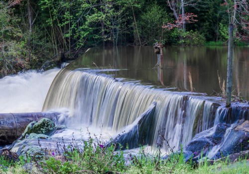A serene waterfall cascades over a rocky ledge into a calm river, surrounded by lush greenery and trees.